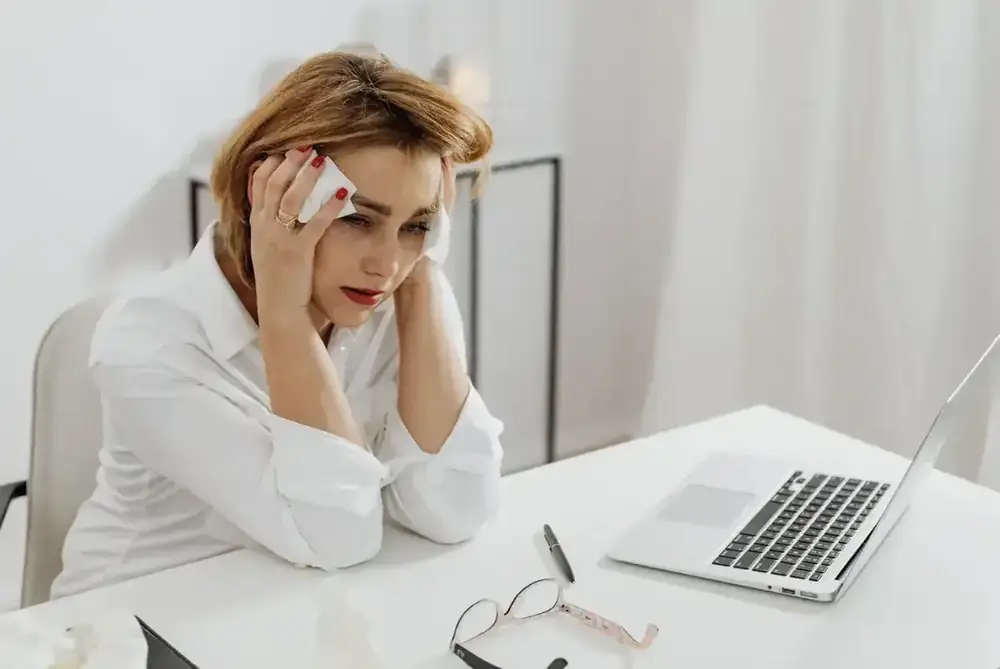 A woman sits at her desk in distress, head in her hands, overwhelmed in front of her laptop.