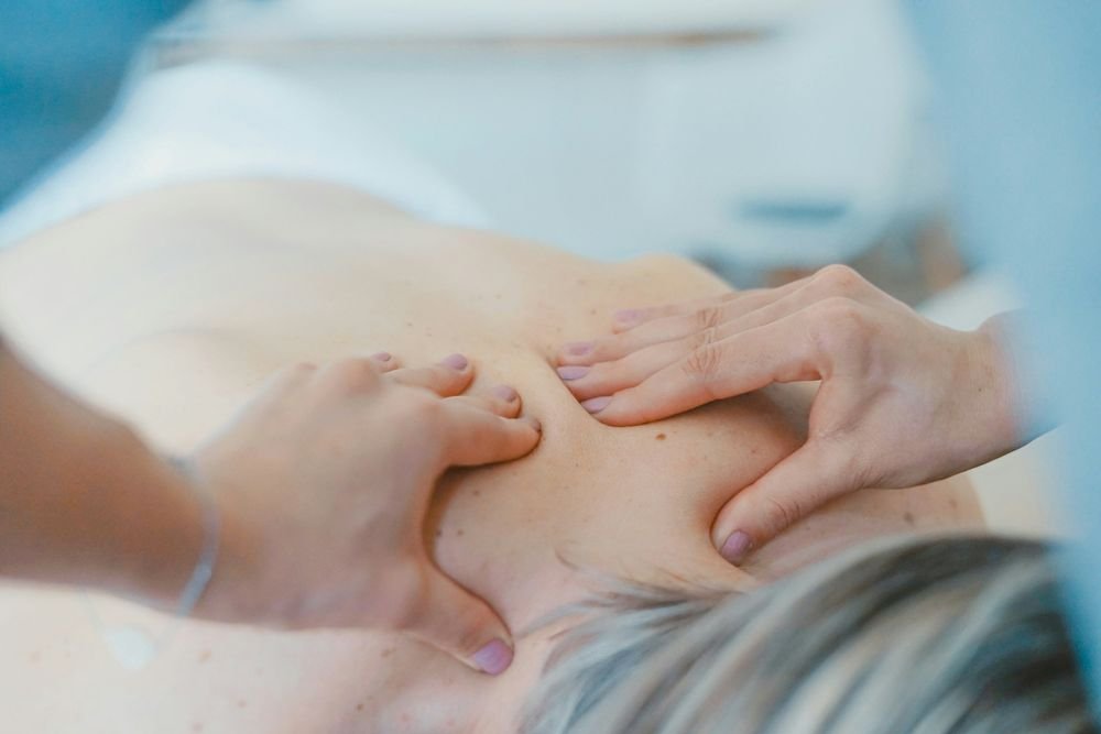 Two hands rest softly on a person’s bare back and shoulder, in soft blue and cream light.