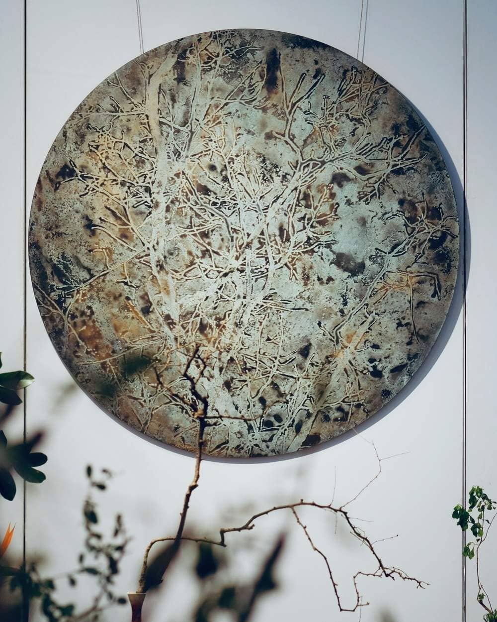 A large round artwork with a pattern like bare branches hangs on a pale wall, with a thin branch in front of it.