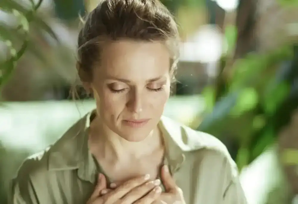 A woman closes her eyes and rests both hands on her chest, breathing calmly among plants.