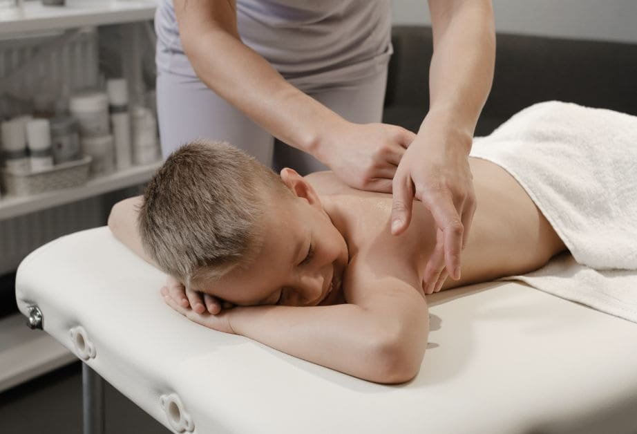A young boy lying relaxed on a treatment couch with his eyes closed while his back is gently massaged.