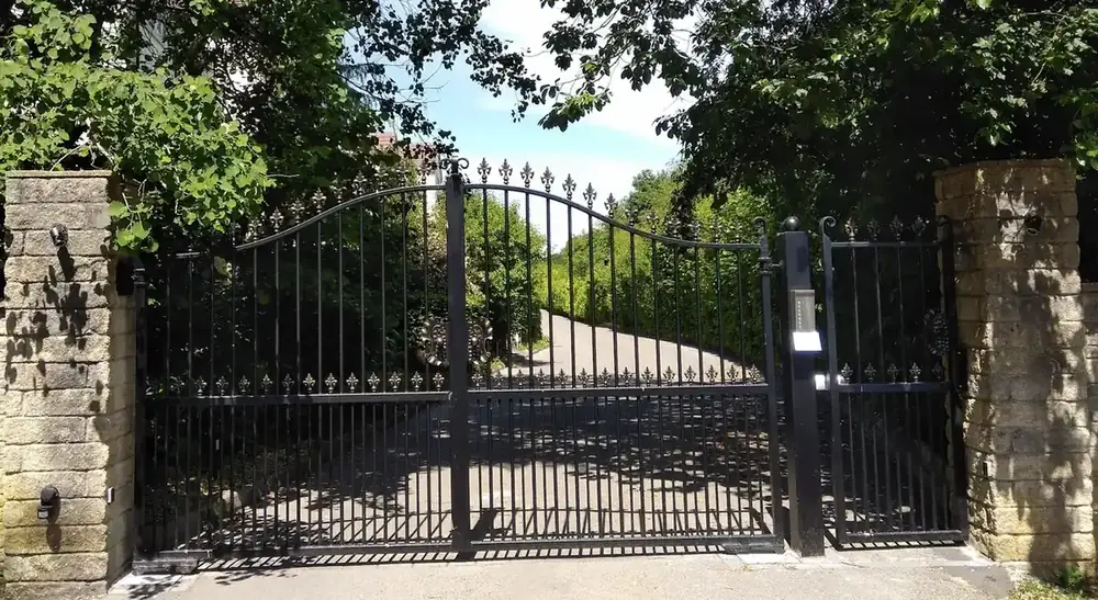 The black iron entrance gates to the clinic, with the intercom on the post to the right and stone pillars and trees either side.