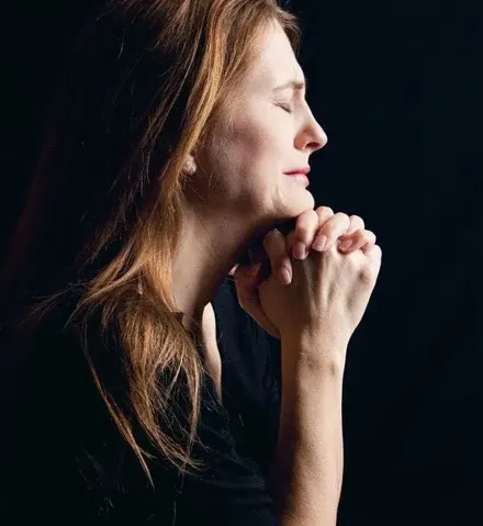 A woman in profile holds her clasped hands to her chin, eyes closed, against a dark background.
