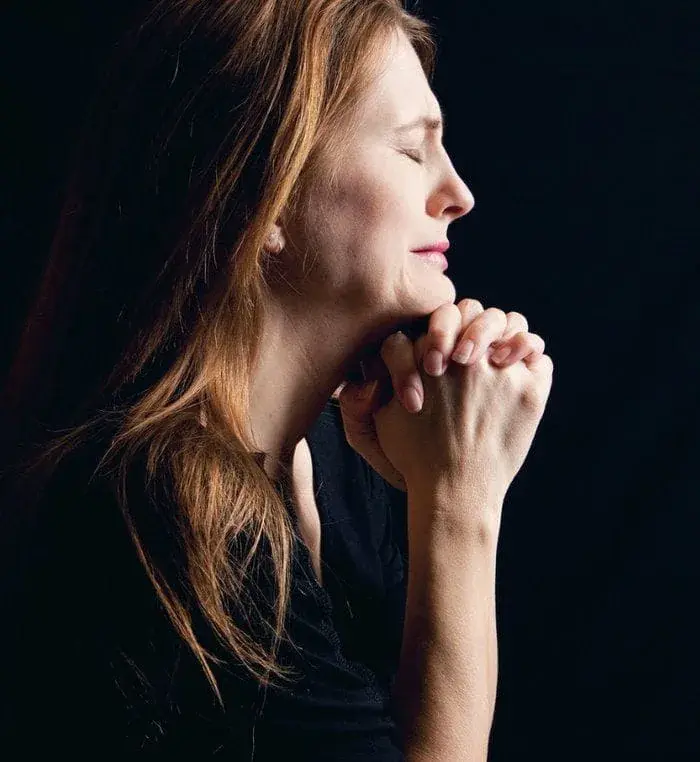 A woman in profile holds her clasped hands to her chin, eyes closed, against a dark background.