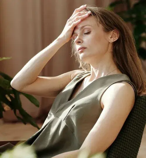 A woman rests her head back in a chair, eyes closed, one hand pressed to her forehead in exhaustion.
