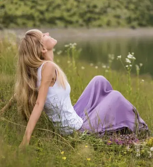 A woman reclines in a meadow by a lake, head tilted back toward the sky, at ease.