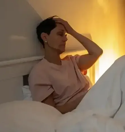 A woman sits up in bed at night by the glow of a lamp, one hand on her head, unable to sleep.
