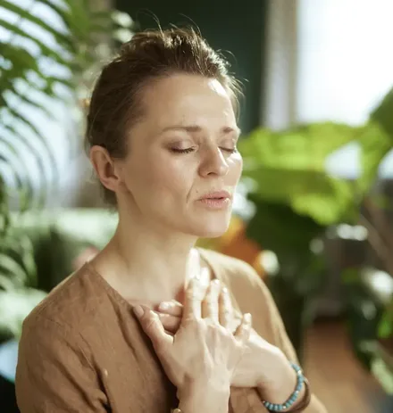 A woman sits with her eyes closed and both hands resting on her chest, breathing slowly.