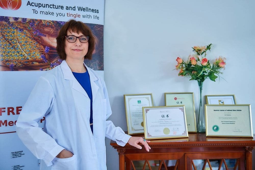 Ela Pękalska in a white clinic coat, smiling in her treatment room beside her framed diplomas in acupuncture, Tui Na and Chinese herbal medicine.
