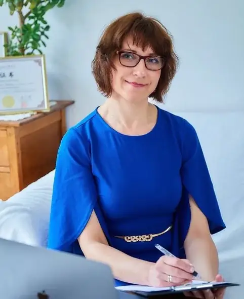 Ela Pękalska smiling in her clinic, in a blue dress, holding a pen and clipboard beside her laptop.
