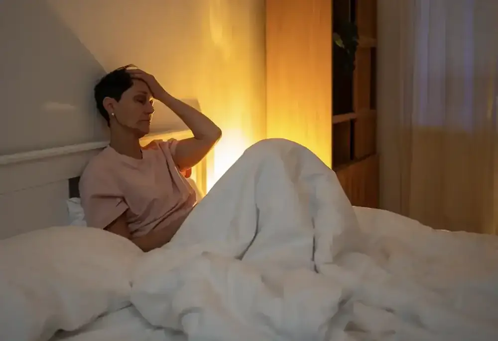 A woman sits up in bed at night, hand pressed to her forehead.