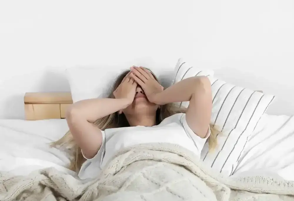 A woman lies in bed with her hands over her face, wrapped in a cosy blanket.