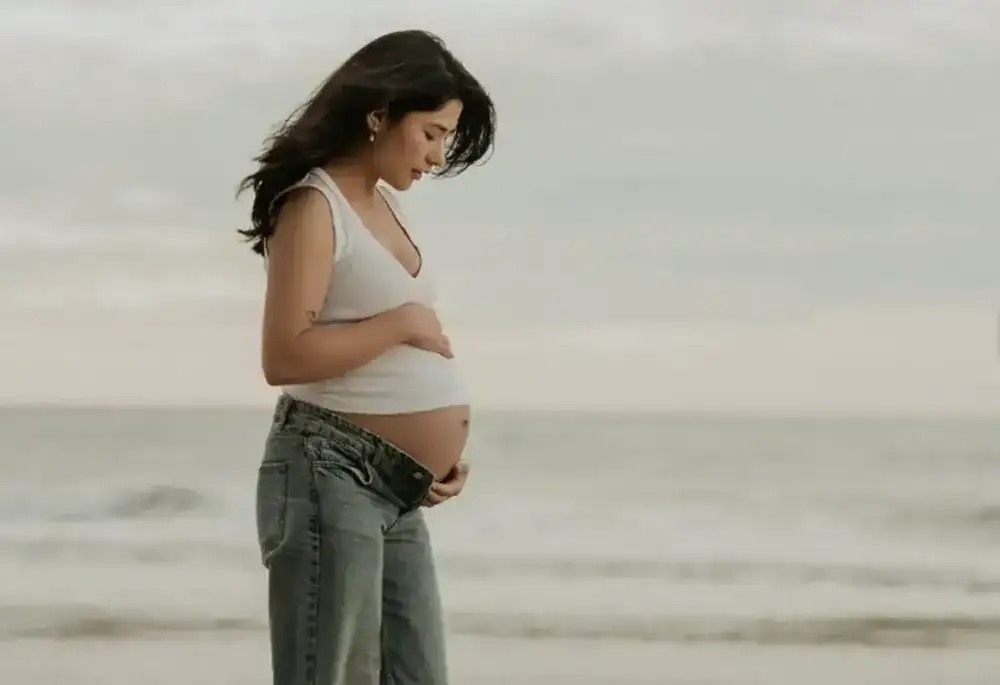 A pregnant woman stands on a beach, looking down at her belly.