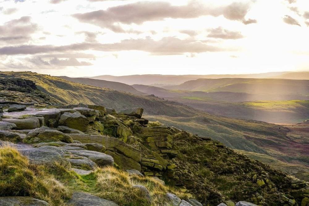Weathered gritstone rocks on a moorland edge, with soft evening light falling across layers of hills.