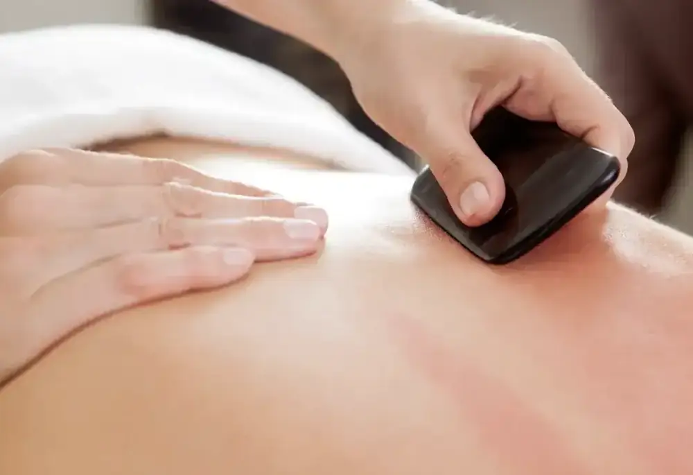 A gua sha tool being drawn across someone's upper back during treatment.
