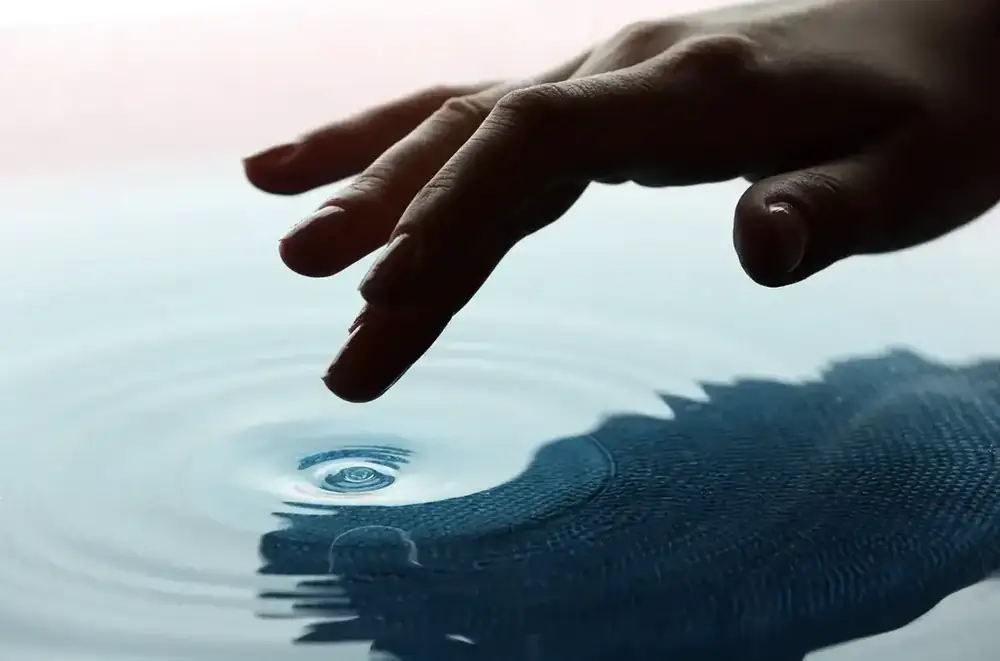 A hand touching still water, sending out soft ripples.