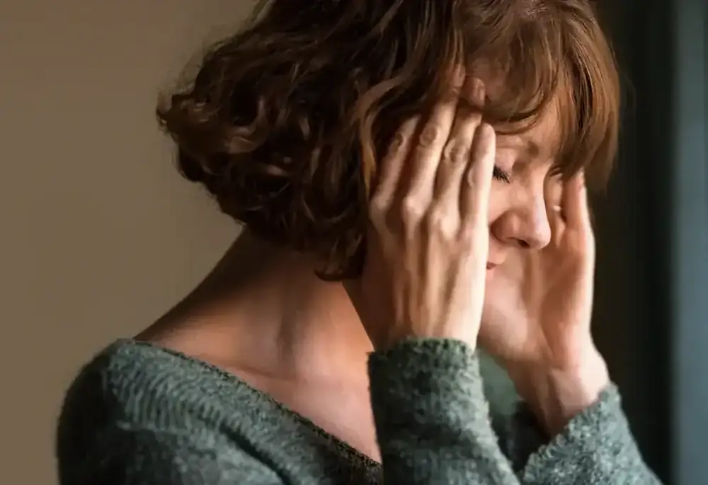A woman presses her hands to her temples, eyes closed, in pain.