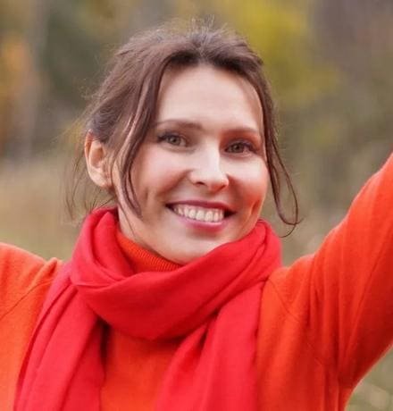 A smiling woman in an orange jumper and red scarf with her arms raised in an autumn field.