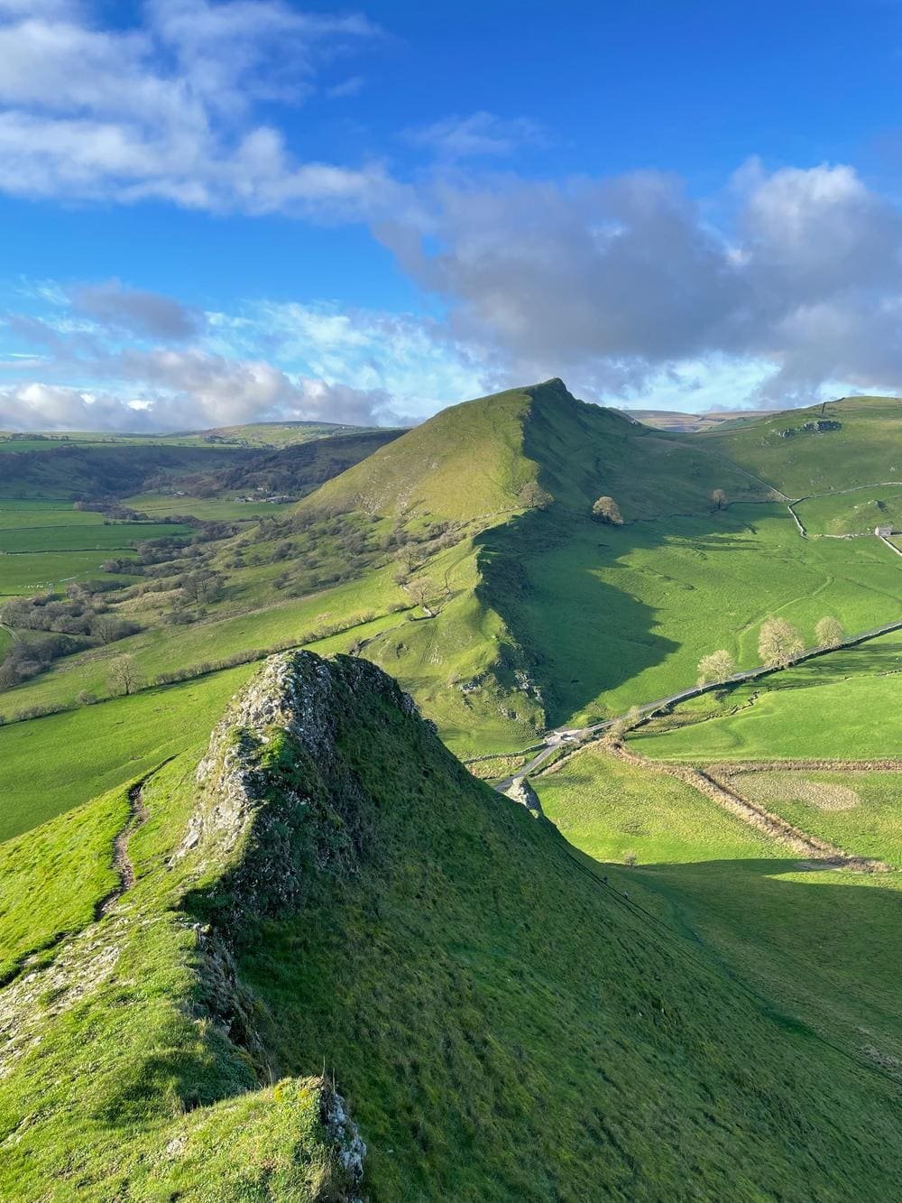 A green limestone ridge running towards a pointed hill under a bright blue sky, with stone walls and fields in the valley.