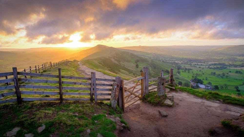 A wooden gate on a stone-flagged ridge path at sunrise, with the hills and green valley fields glowing below.
