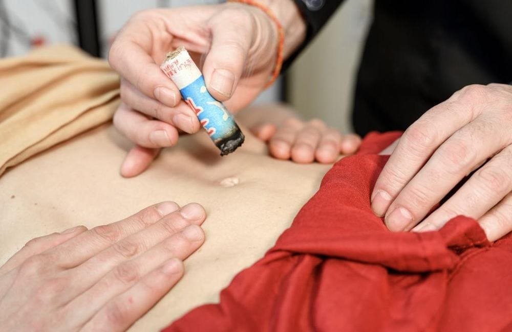 A lit moxa roll held close to the abdomen beside the navel, while the practitioner's other hand rests on the belly.