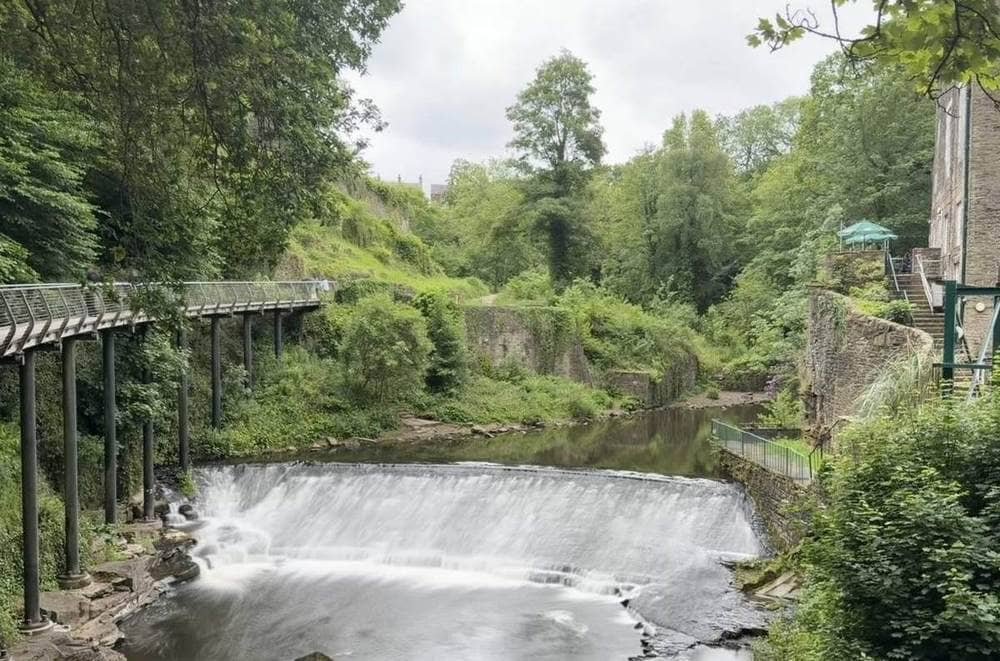 The Millennium Walkway on tall pillars beside a curved weir on the river in the Torrs gorge at New Mills, with a stone mill and trees.