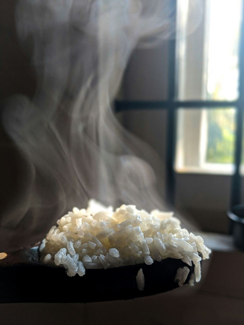 Steam rising from freshly cooked white rice on a dark wooden spoon, with soft light from a window behind.