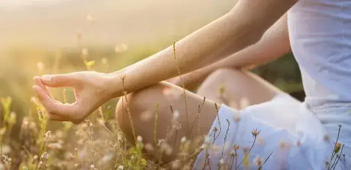 A woman sits cross-legged outdoors in a meadow at golden hour, one hand resting in a meditation mudra.