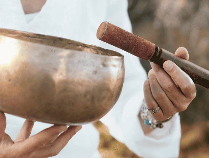 Hands holding a brass singing bowl and a wooden mallet outdoors.