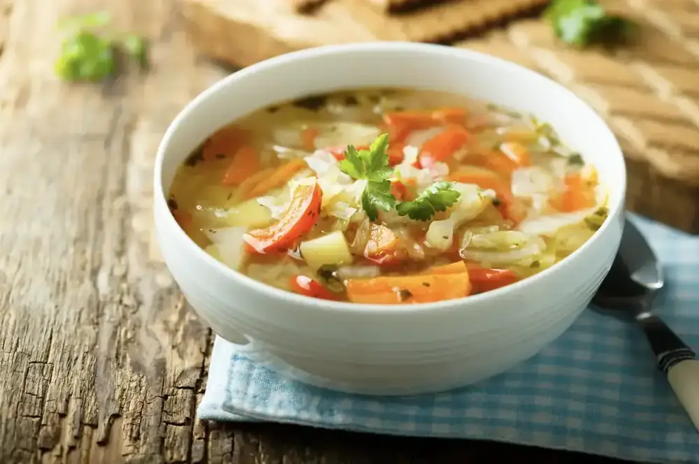 A bowl of warming vegetable soup with carrot, cabbage and coriander, set on a wooden table.