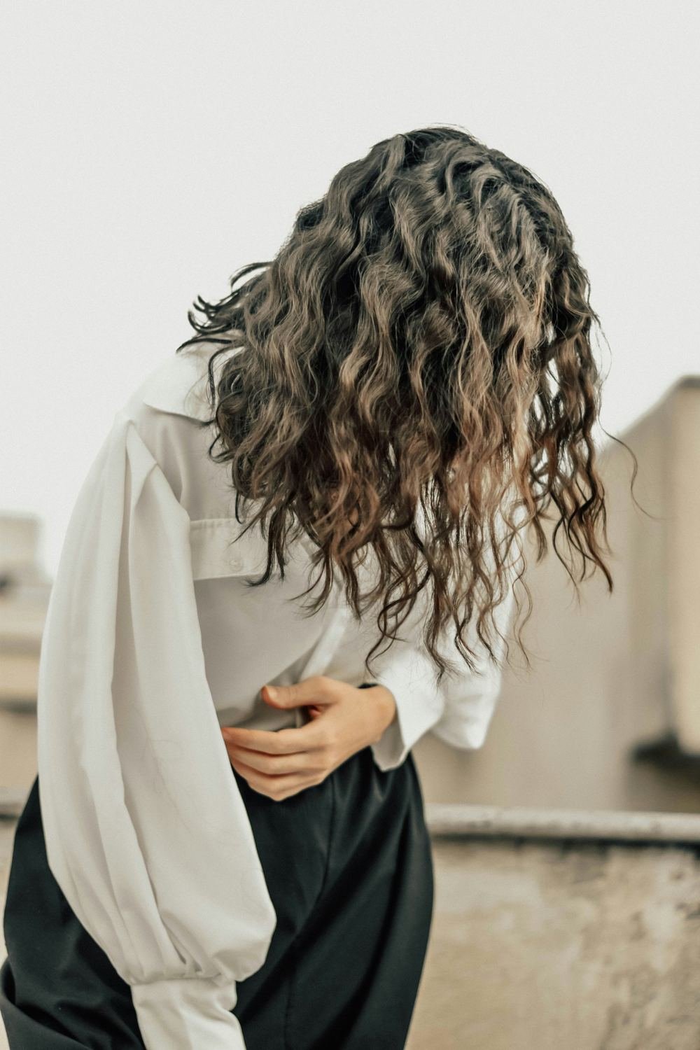 A woman in a white shirt bends forward with her long curly hair falling over her face, one hand pressed to her stomach.