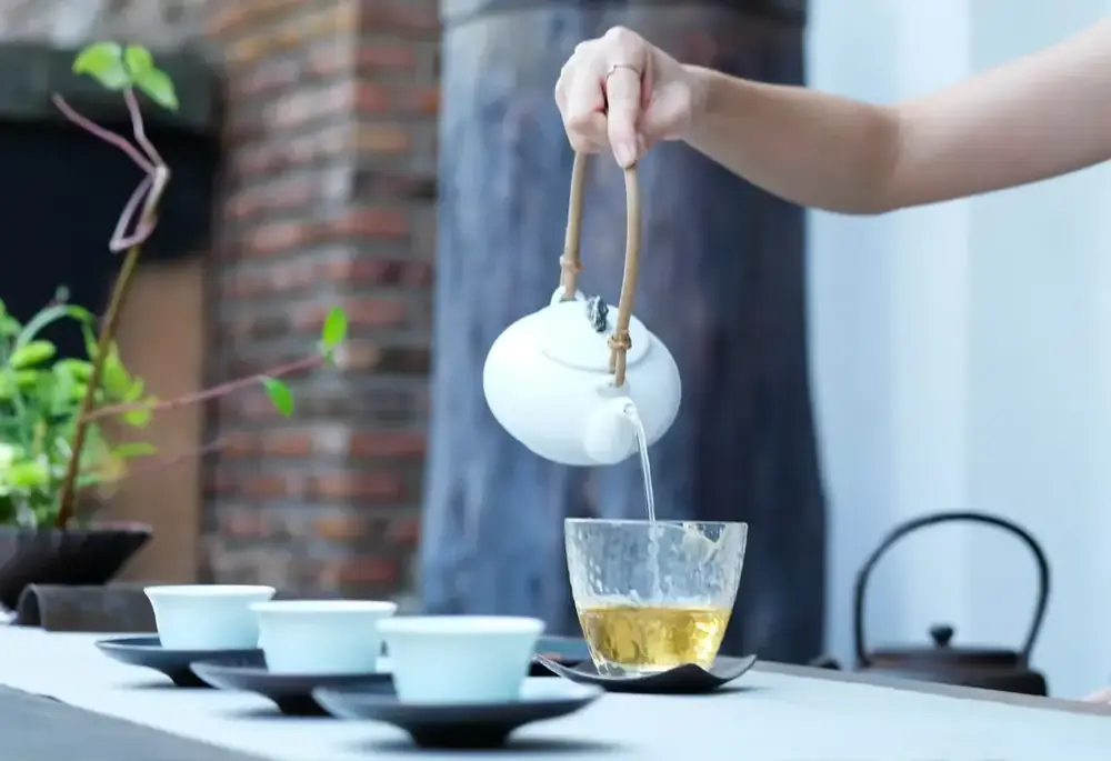 A hand pours pale tea from a white teapot into a glass cup, with small porcelain cups set out on the table.