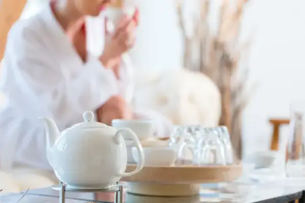 A woman relaxes with a cup of tea in a robe, with a white teapot and glasses set out nearby.