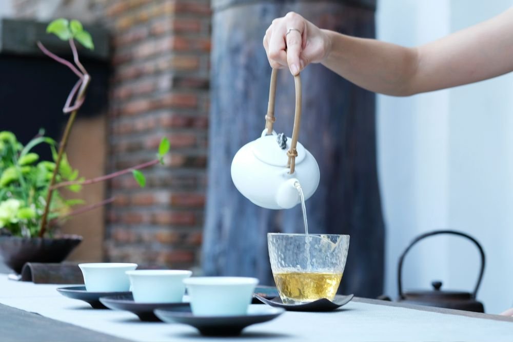 A hand pours tea from a white teapot into a glass cup, with small white cups and a green plant on the table.