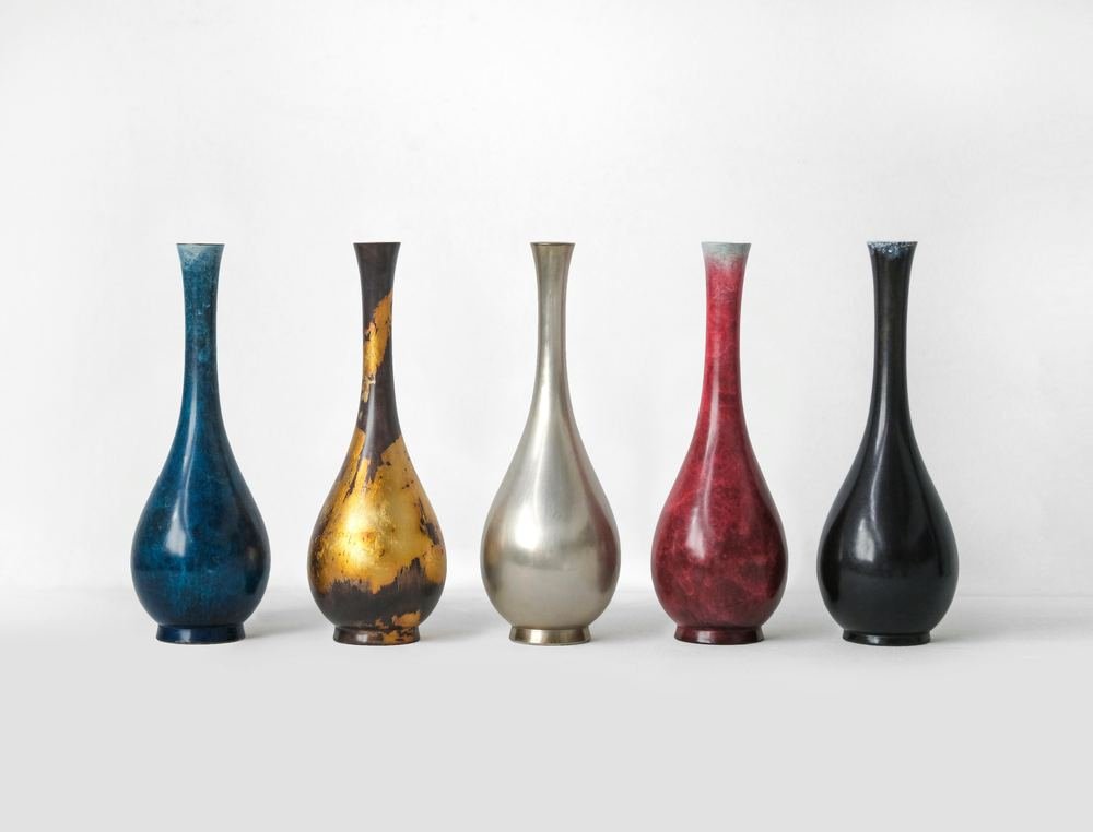 Five slender bronze vases in blue, gold leaf, silver, red and black, standing in a row against a pale wall.