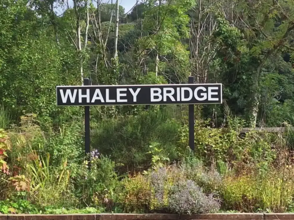 The Whaley Bridge station sign, framed by trees and a garden border.