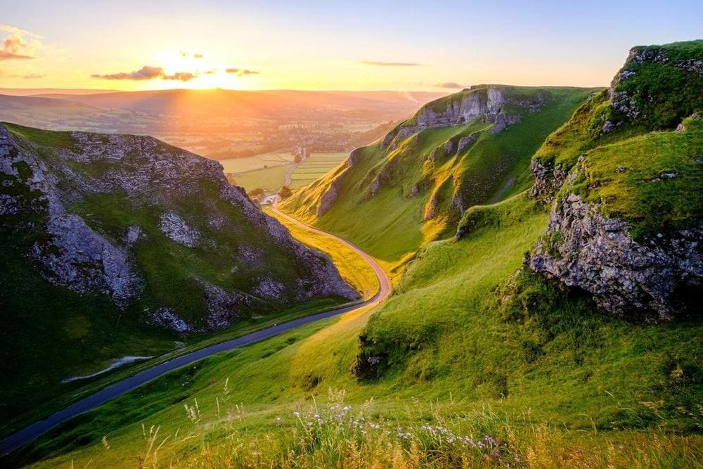 A narrow road winding through a steep green limestone pass at sunset, with the valley and village glowing below.