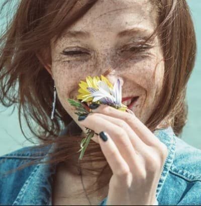 A smiling woman with freckles and windswept red hair, eyes closed, holding a small bunch of flowers to her face.