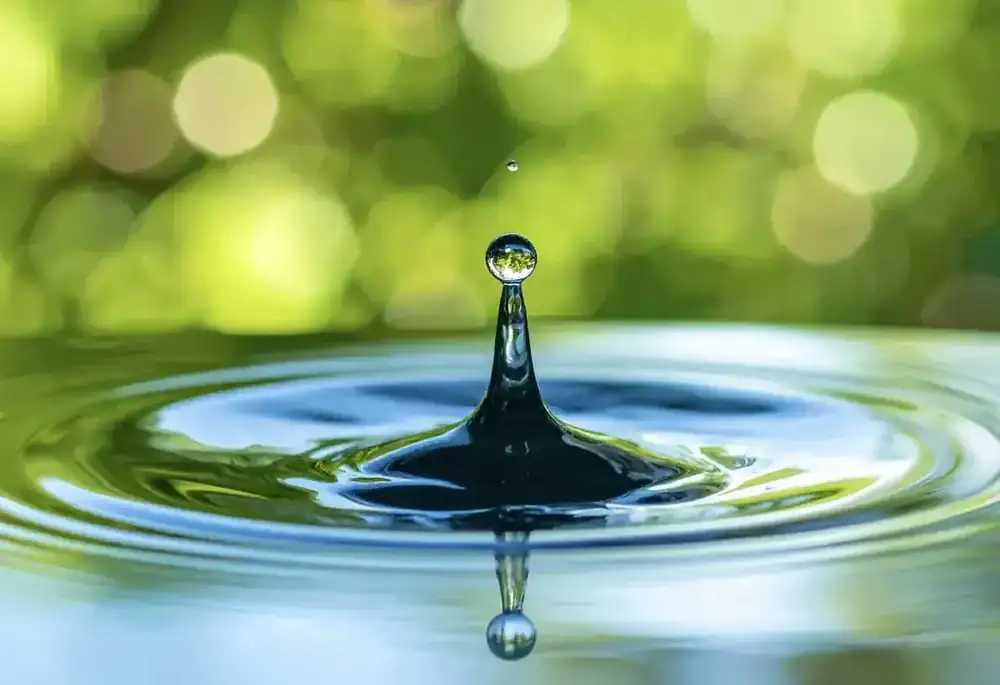 A single water droplet rising from a still pool, with soft green light behind it and ripples spreading outwards.