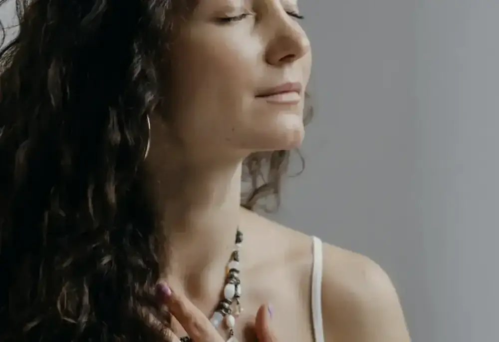 A woman with her eyes closed rests her hand gently at her chest, serene.