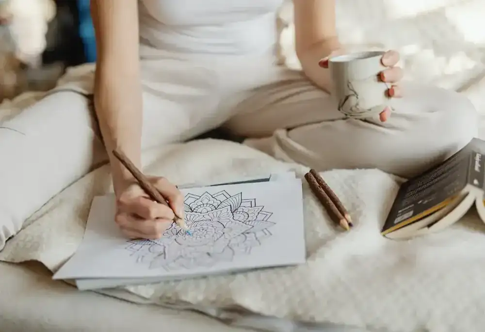 A woman colours a mandala while holding a cup of tea in a cosy nook.