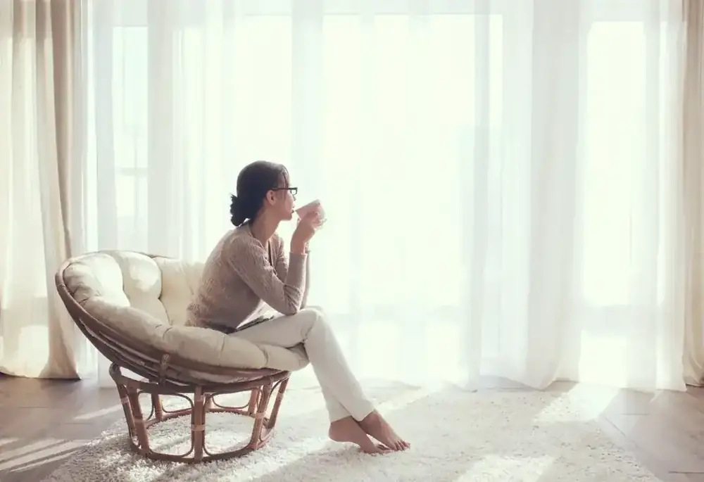 A woman sits in a papasan chair sipping tea beside a sunlit window.